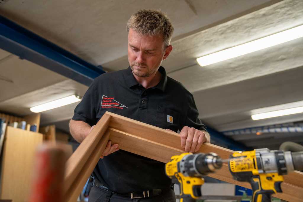 En tømrer i sort poloshirt holder et trævinduesramme i et værksted.